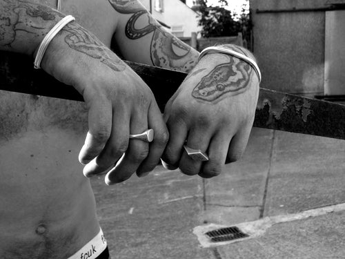 Tattooed male hands and arms rest on a steel handrail. They are wearing chunky silver signets, a bangle and chain bracelets. the background is a bare concrete yard.