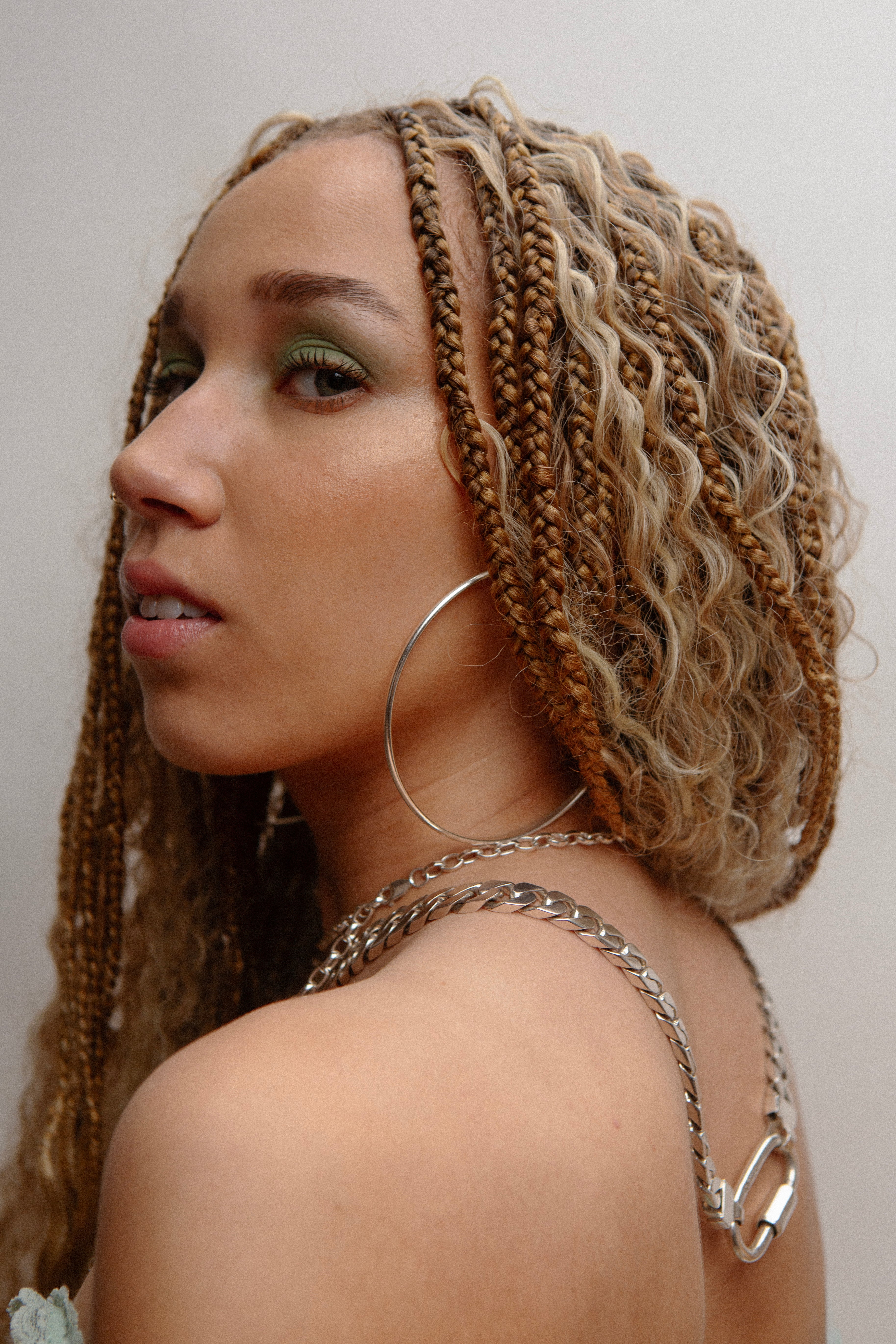 Woman with braids and wavy hair wears large silver hoop earrings and two chunky silver chains. She looks sideways at the camera.