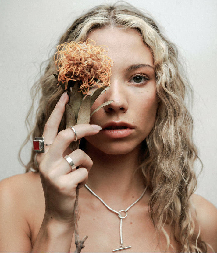 Woman with a flower near her eye against a plain background wears sustainably made silver jewellery including a red coral ring, silver rings and a silver chian necklace